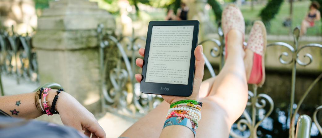 A tattooed woman wearing bracelets, with her feet up against a metal fence, reading on a Kobo ereader